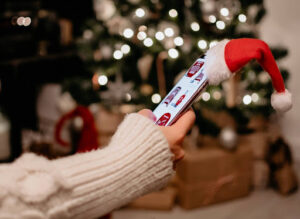 A hand holds a cell phone with a Christmas hat on top of it. A lit Christmas tree and gifts are blurred in the background